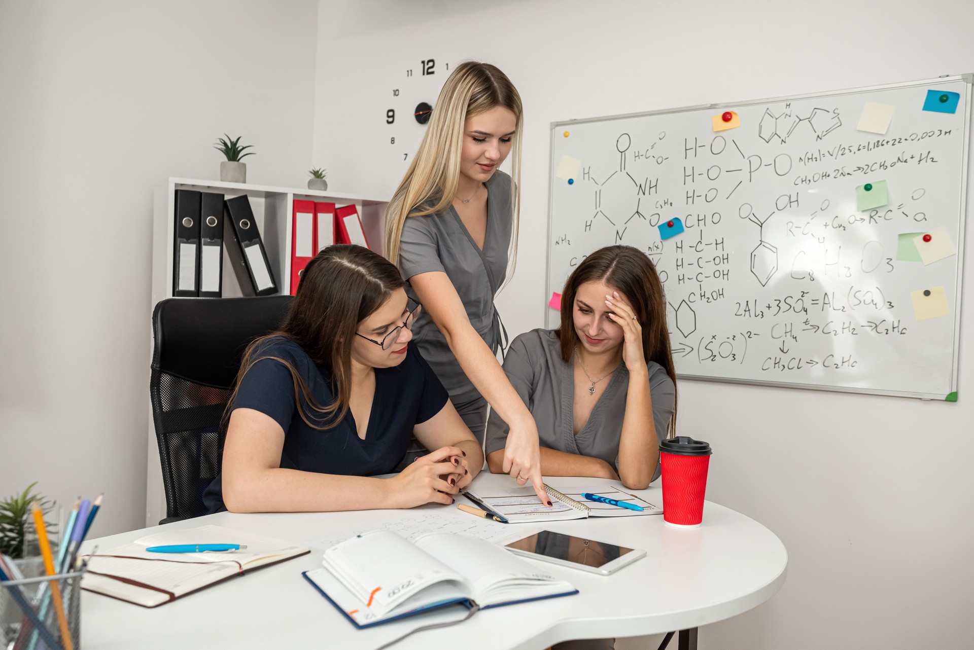 Teamwork and discussion between three women, chemistry formulas written on blackboard.