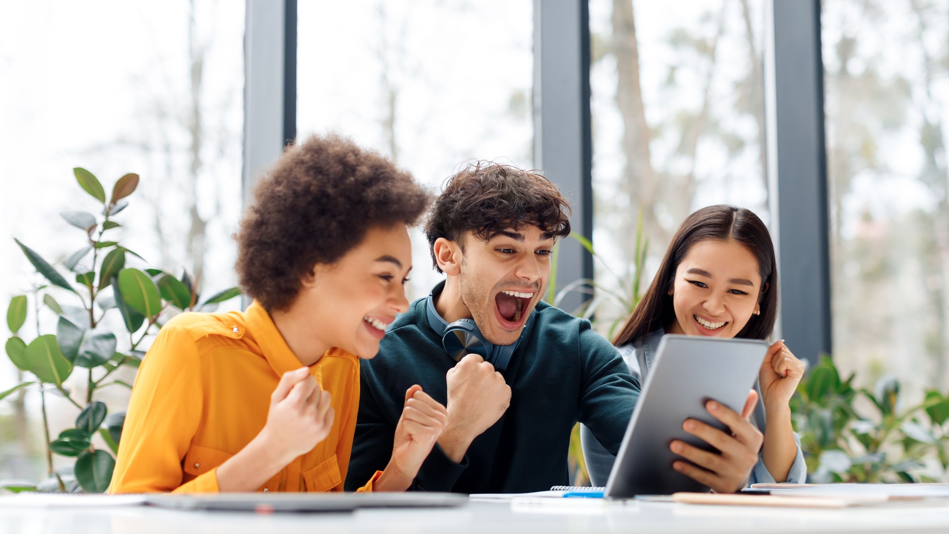 Overjoyed multiracial classmates looking at digital tablet screen, shouting yes and clenching fists in classroom